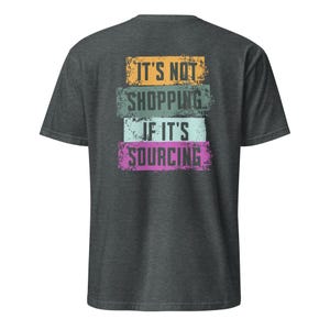 May include: Dark gray t-shirt with the text "IT'S NOT SHOPPING IF IT'S SOURCING" in a layered design. The text is in orange, teal, light blue, and pink. The shirt is a short-sleeved crew neck.