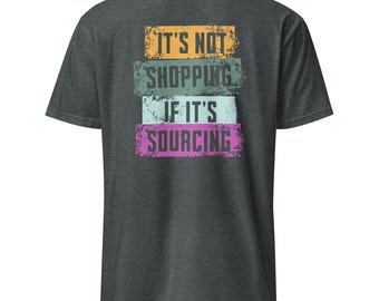 May include: Dark gray t-shirt with the text "IT'S NOT SHOPPING IF IT'S SOURCING" in a layered design. The text is in orange, teal, light blue, and pink. The shirt is a short-sleeved crew neck.