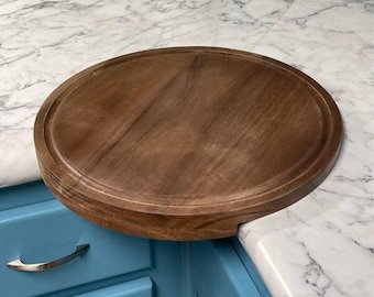 May include: A round wooden cutting board with a raised edge, mounted on a corner of a countertop. The board is made of dark brown wood and has a natural finish.
