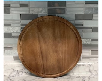 May include: A round wooden cutting board with a natural brown finish. The board has a slightly raised edge around the perimeter.