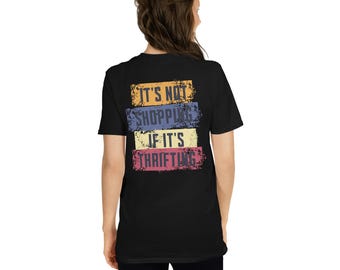 Puede incluir: Camiseta negra con el texto "IT'S NOT SHOPPING IF IT'S THRIFTING" en un diseño colorido y desgastado. El texto está dispuesto en cuatro bloques horizontales de color: naranja, azul, amarillo y rosa. La camiseta es de manga corta.