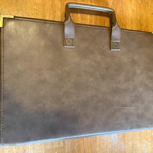 May include: A brown briefcase with a handle and gold-colored corner accents. The briefcase is rectangular and appears to be made of a leather-like material. The handle is attached with two straps.