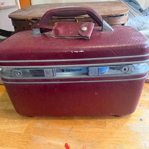 May include: A maroon hardshell Samsonite train case with a textured exterior, a top handle, and a matching luggage tag. The front features a silver-toned metal trim with two integrated locking latches and the Samsonite brand name printed in black.