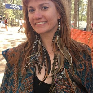 May include: A woman with long, red hair wearing a blue and orange paisley shawl with fringe. She is wearing a black tank top and has long feather earrings.