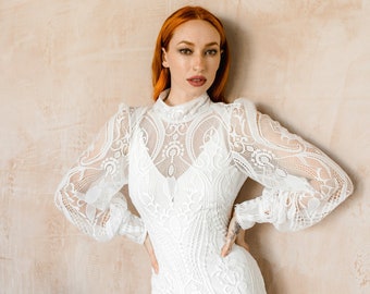May include: A white lace wedding dress with long sleeves and a high neckline. The dress has a fitted bodice and a flowing skirt with a slit in the front.