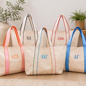 Puede incluir: Cinco bolsos de lona color hueso con asas de borde festoneado en naranja, negro, multicolor, rojo y azul. Cada bolso presenta letras griegas bordadas: ΑΓΔ, ΧΩ, ΚΚΓ, ΠΒΦ y ΔΓ centradas en el panel frontal de la tela.