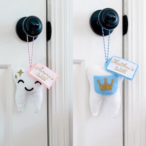 May include: Two white felt tooth-shaped pouches hanging from black door knobs. The left pouch has a smiling face, gold stars, and a pink tag reading Aurora's tooth. The right pouch has a blue pocket with a gold crown and a blue tag reading Matthew's tooth.
