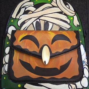 Könnte beinhalten: Ein Halloween-Rucksack mit einem Mumien-Design auf der Oberseite und einem Kürbis-Laternen-Motiv auf der Frontklappe. Die Tasche hat einen grünen Hintergrund mit weißen und schwarzen Akzenten und einen goldfarbenen Verschluss.