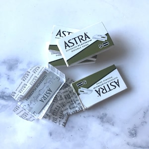 May include: A close-up of a pack of Astra Superior Platinum Double Edge razor blades. The pack is green and white with the brand name and product description printed on it. The blades are individually wrapped in paper.