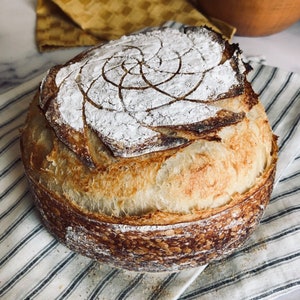May include: A freshly baked loaf of sourdough bread with a spiral design on top. The bread has a golden brown crust and is sitting on a white and blue striped kitchen towel.