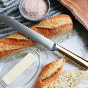 Puede incluir: Primer plano de una encimera de cocina con una barra de pan francés, un cuchillo, un bol de sal rosa y un plato de mantequilla. El pan está cortado y listo para ser untado.