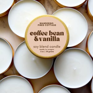 May include: Overhead shot of several small, round soy blend candles with white wax and gold-colored metal containers. The central candle has a label that reads "coffee bean & vanilla" and "soy blend candle". The label also states "made in oregon" and "2 oz | 55 grams".