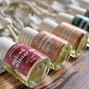 May include: A row of small 8 ml glass car diffuser bottles with wooden caps and hanging cords. Each bottle features a colored label with text including Spiced Winter Tea, Toasted Pumpkin Spice, and other scents, arranged in a diagonal line.