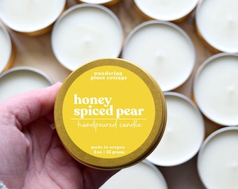 May include: A gold tin candle with a yellow label that reads "wandering pines cottage honey spiced pear handpoured candle made in oregon 2 oz | 55 grams"