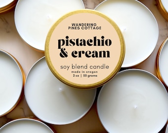 May include: A round gold metal tin containing a white soy blend candle with a beige label that reads Wandering Pines Cottage, Pistachio & Cream, soy blend candle, made in Oregon, 2 oz | 55 grams. The candle features a centered cotton wick.