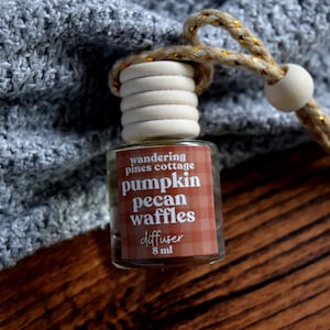 May include: A small glass diffuser bottle with a wooden cap and a decorative rope. The label reads "wandering pines cottage pumpkin pecan waffles diffuser 8 ml". The background includes a gray knitted fabric and a wooden surface.