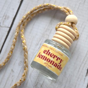 May include: A small glass diffuser bottle with a wooden top and a braided cord. The label reads "cherry lemonade diffuser 8 ml." The cord is beige with gold accents. The bottle is on a light wood surface.