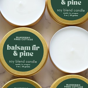 May include: Round gold metal tins containing white soy blend candles. The dark green labels feature the text Wandering Pines Cottage, balsam fir & pine, soy blend candle, made in oregon, 2 oz | 55 grams. The candles have a single centered wick.