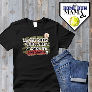 Könnte beinhalten: Schwarzes T-Shirt mit einer grünen und roten Grafik, auf der "Ich bin die Mama, die nicht neben dem Spielveränderer sitzen kann!!" steht, sowie einem Baseball-Motiv. Das T-Shirt liegt auf einem Holzgrund mit einer blauen Jeans.