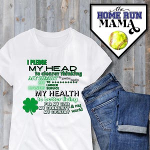 Può includere: Maglietta bianca con testo verde che recita "Prometto la mia testa a un pensiero più chiaro, il mio cuore a una maggiore lealtà, le mie mani a un servizio più ampio, la mia salute a una vita migliore, per il mio club, la mia comunità e il mio mondo". Un trifoglio a quattro foglie verde si trova a sinistra del testo. La maglietta è su una superficie di legno con un paio di jeans blu piegati sopra.