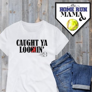 May include: White t-shirt with black text that reads "CAUGHT YA LOOKIN'" with a red "K" and a baseball graphic. The t-shirt is on a wood background with a blue and white baseball home plate sign that reads "The Home Run Mama".