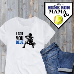 Może przedstawiać: Biała koszulka z czarną sylwetką baseballisty łapiącego piłkę. Tekst "I got you blue" jest nadrukowany na koszulce czarnym i niebieskim kolorem.
