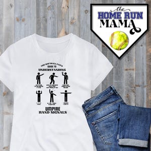 May include: White t-shirt with a black graphic of a softball umpire's hand signals. The graphic includes the text "The Softball Fan's Guide to Understanding Umpire Hand Signals".