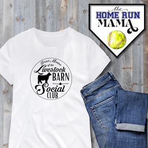 Può includere: Maglietta bianca con una grafica nera che dice "Proud Member of the Livestock Barn Social Club". La grafica presenta una silhouette di una mucca. La maglietta è appoggiata su una superficie di legno accanto a un paio di jeans blu.