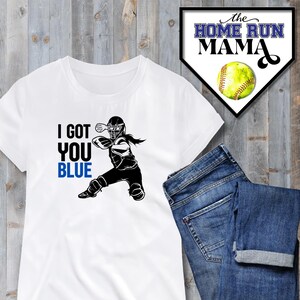 Może przedstawiać: Biała koszulka z czarną sylwetką gracza softballa łapiącego piłkę. Tekst "I GOT YOU BLUE" jest nadrukowany na koszulce na czarno i niebiesko.