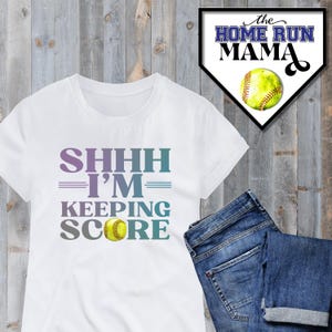 May include: White t-shirt with the text "SHHHH I'M KEEPING SCORE" in a gradient of blue and purple. A yellow softball is centred below the text.