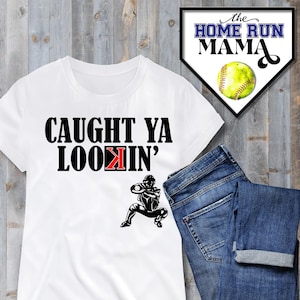 May include: White t-shirt with black text that reads "CAUGHT YA LOOKIN'" and a black silhouette of a softball player catching a ball. The text "K" in "LOOKIN'" is red.