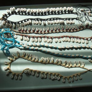 May include: Four handmade necklaces featuring white cowrie shells strung on cord. The top necklace uses black cord with black beads, the second uses brown cord with brown beads, the third uses bright blue cord, and the bottom uses tan cord with tan beads.
