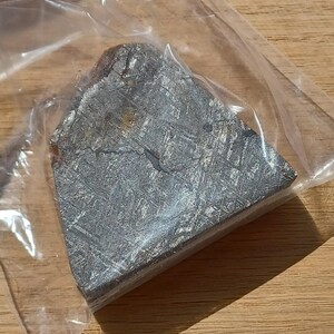 May include: A triangular-shaped iron meteorite fragment with a dark, metallic gray surface featuring a distinct Widmanstätten pattern of intersecting crystalline lines. The specimen is encased in a clear, thin plastic storage bag.