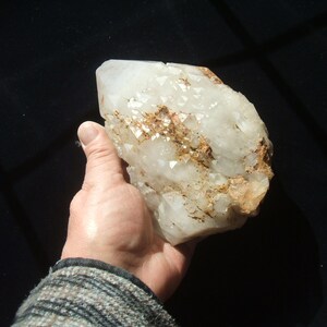 May include: A large, raw quartz crystal with a milky white appearance and areas of golden-brown mineral deposits. The crystal is held in a hand, showcasing its size and natural texture. The background is a dark, solid color.