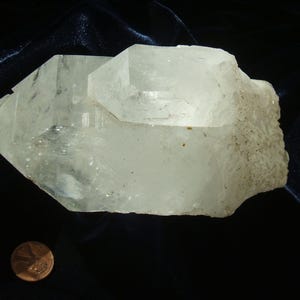 May include: A large, clear quartz crystal cluster with multiple points. The crystal is translucent and has a slightly cloudy appearance. The crystal is sitting on a dark background.