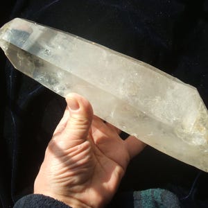 May include: A large, clear quartz crystal with a pointed tip and a rough, natural texture. The crystal is held in a hand, showcasing its size and clarity. The crystal is a light, translucent color.