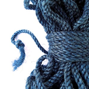 May include: A coil of thick, dark blue twisted rope made of natural fibers. The rope is wound tightly in a cylindrical shape with a central binding section. A loose, frayed end of the rope extends outward from the main coil, showing the texture.