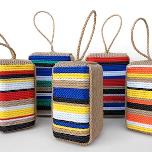 May include: Five colorful doorstops made of woven rope with a loop handle. The doorstops are striped with different colors, including red, yellow, blue, green, black, and white.