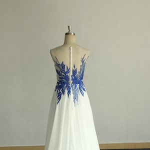 May include: A white chiffon wedding dress with a blue lace applique on the back. The dress has a scoop neckline and a flowing skirt.