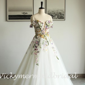 Puede incluir: Un vestido de novia blanco con bordado floral. El corpiño y la falda con hombros descubiertos están adornados con flores de colores. El vestido se exhibe en un maniquí. El texto "Vickymermiadbridal" es visible.