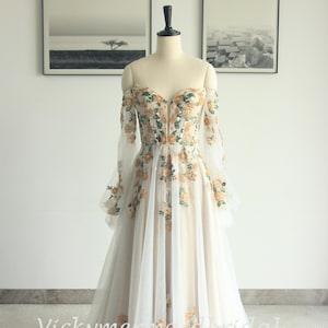 May include: Off-the-shoulder wedding dress with floral embroidery. The ivory gown features long, sheer sleeves and a sweetheart neckline. The bodice and skirt are adorned with orange and green floral patterns. The dress is displayed on a mannequin.