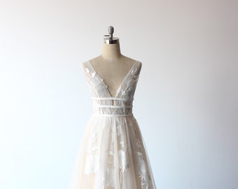 May include: A white wedding dress with a plunging V-neckline and delicate floral embroidery. The dress has a fitted bodice and a flowing A-line skirt. The dress is on a mannequin against a white background.