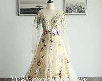 May include: A cream-colored, floor-length gown with a deep V-neck and long, sheer sleeves. The dress is adorned with colorful floral embroidery in shades of red, purple, yellow, and white, with green foliage. The text "Vickymermad" is visible on the dress.