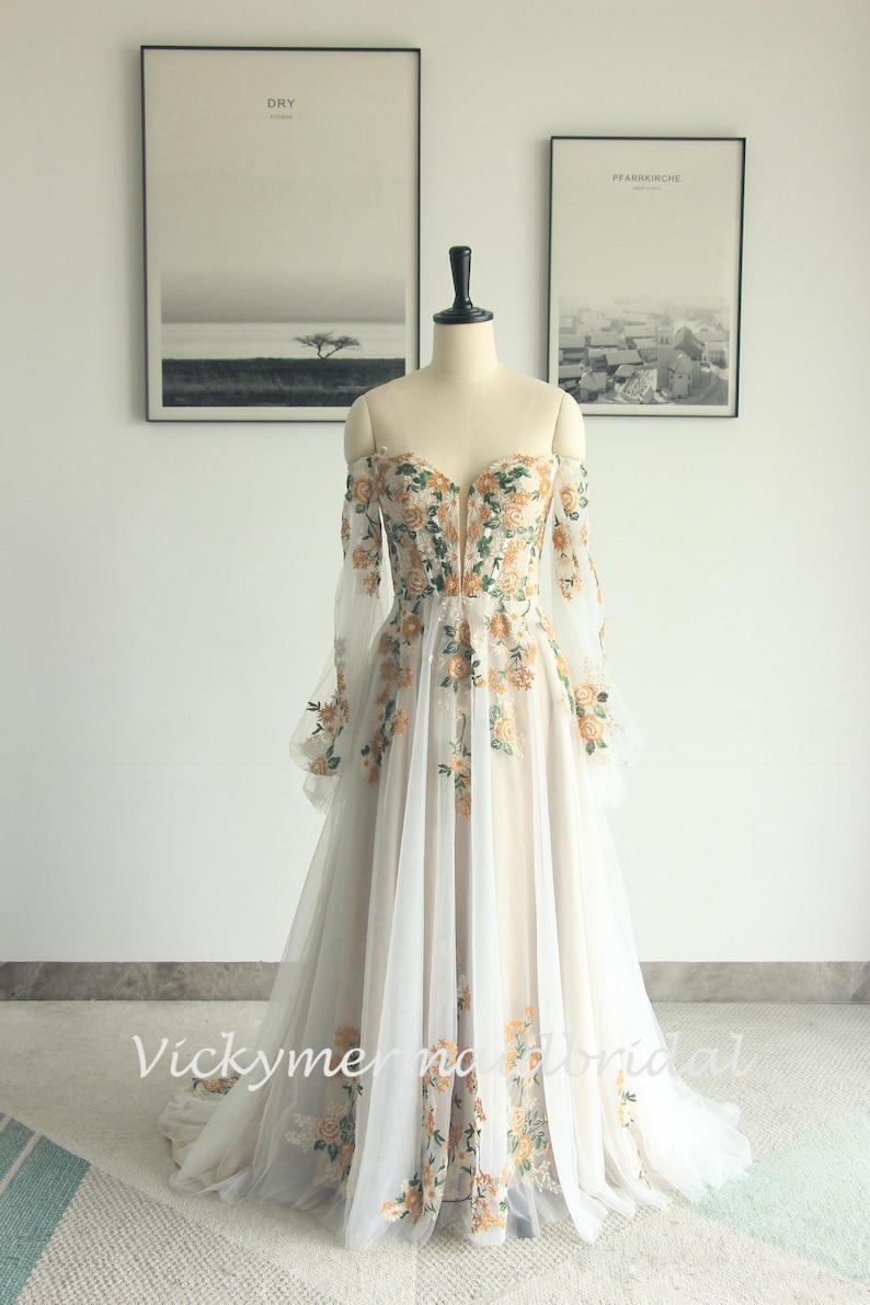 May include: Off-the-shoulder wedding dress with floral embroidery. The ivory gown features long, sheer sleeves and a sweetheart neckline. The bodice and skirt are adorned with orange and green floral patterns. The dress is displayed on a mannequin.