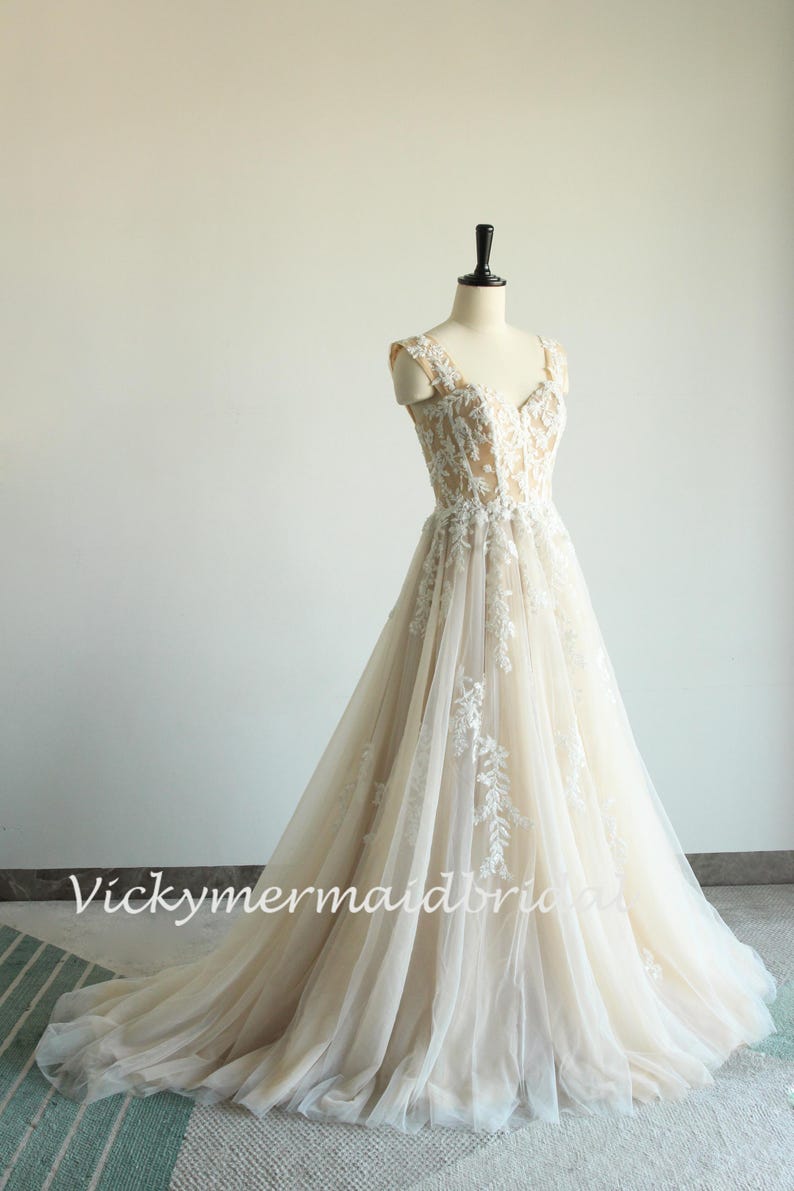 May include: A sleeveless, ivory wedding dress with a sweetheart neckline and delicate floral lace embroidery. The dress has a flowing A-line skirt and a sheer tulle overlay.