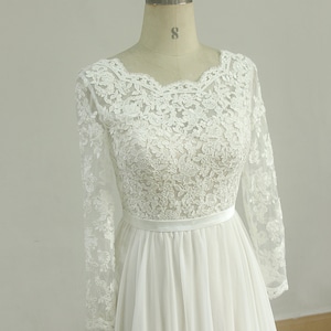 May include: A white long-sleeved lace wedding dress with a scoop neckline and a chiffon skirt. The dress has a satin sash at the waist.