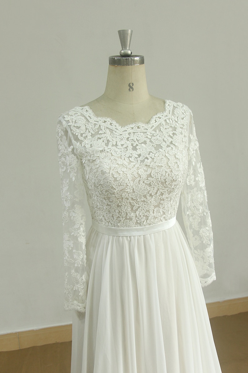 May include: A white long-sleeved lace wedding dress with a scoop neckline and a chiffon skirt. The dress has a satin sash at the waist.