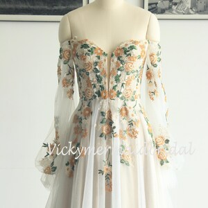 May include: Off-the-shoulder ivory wedding dress with floral embroidery in orange and green. The long sleeves are sheer, and the bodice features a deep V-neck. The skirt is a flowing, layered design. The text "Vickymerybridal" is visible.