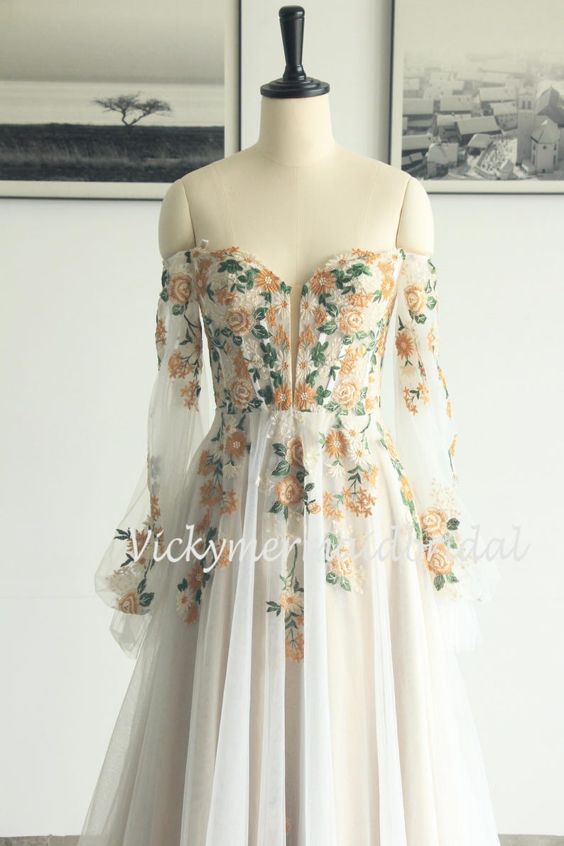 May include: Off-the-shoulder ivory wedding dress with floral embroidery in orange and green. The long sleeves are sheer, and the bodice features a deep V-neck. The skirt is a flowing, layered design. The text "Vickymerybridal" is visible.