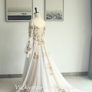 May include: Off-the-shoulder wedding gown with floral embroidery. The ivory dress features long, sheer sleeves and a flowing train. The bodice and train are adorned with orange and green floral accents. The dress is displayed on a mannequin.